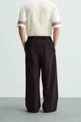 Pantalón marrón oscuro de corte ancho, confeccionado en mezcla de algodón y lino. Presenta cintura elástica, bolsillos laterales y traseros, pliegues frontales y cierre con cremallera y botón.