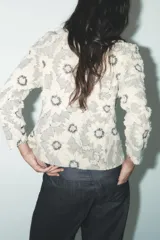 Blazer color crudo con estampado de flores negras, cuello solapa con muescas, manga larga y cierre frontal con botones negros.