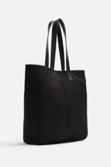 Bolso tipo shopper negro fabricado en tejido de algodón con asas de piel. Cuenta con dos bolsillos laterales exteriores y un bolsillo interno.