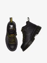Botín de cuero negro Dr. Martens modelo Woodard, con cuello acolchado, cordones de tipo excursionista en amarillo y negro, suela acanalada Club Wedge con pespunte negro y tirador trasero con logo.