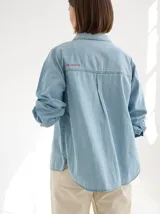 Camisa de denim azul con pespuntes blancos, corte crop y cierre frontal con botones. Tiene cuello clásico, mangas cortas y un bolsillo de parche en el pecho. Se ata en la parte delantera.
