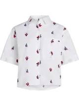 Camisa blanca de manga corta con cuello camisero, cierre de botones frontal y bolsillo de parche en el pecho. Presenta un bordado floral integral en tonos rojo, azul y negro.