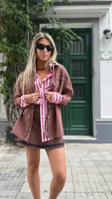 Camisa de denim color chocolate con botones dorados, corte recto, cuello clásico y mangas largas con puños abotonados. Presenta detalles de costuras en contraste y bolsillos frontales.