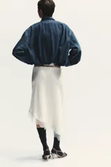 Chaqueta corta de denim azul con cuello alto, manga larga con hombros caídos, bolsillos laterales, bajo con elástico y cierre frontal con cremallera y botones ocultos.
