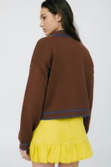 Cárdigan tejido en 50% algodón y 50% acrílico, color crema, con pespuntes tejidos en contraste color borgoña y bordado de limón en el pecho. Tiene cuello con volados, cierre frontal con botones y corte relajado.