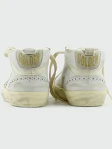 Zapatillas de caña media estilo deportivo, color blanco, con efecto desgastado en la suela. Presentan el logo estrella lateral cubierto de purpurina plateada y detalles perforados en el cuero. Llevan cordones blancos y la inscripción "GGDB MID/STAR" en el lateral.