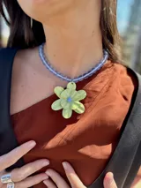 Collar corto con cuentas de vidrio color lila y dije de flor de seis pétalos color verde lima con centro celeste. Cierre con cadena de acero quirúrgico y ganchito para regular el largo.
