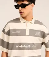 Camisa polo oversized con cuello abotonado, mangas cortas y diseño a rayas horizontales en tonos crema y gris. Presenta estampados con texto en el pecho.