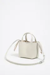 Bolso de mano mini de piel vacuna color blanco roto, con textura granulada. Presenta asas superiores y cordones ajustables laterales. El logo metálico de Bimba y Lola está grabado en el frente.