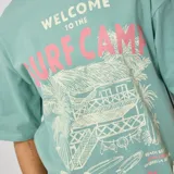 Remera verde con estampado de cabaña de playa, palmeras y tablas de surf.
