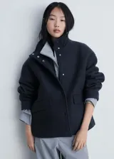 Chaqueta corta de lana color gris oscuro, con cuello alto, cierre frontal con botones a presión, bolsillos laterales y forro interior.