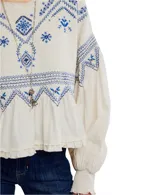 Blusa color crema con bordados azules, mangas largas abullonadas con puños elásticos y cuello redondo con detalle de volados.