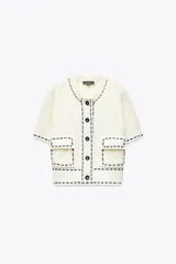 Chaqueta color crudo de tejido de punto con cuello redondo y manga corta. Tiene bolsillos delanteros de plastrón con solapa, detalle de pespuntes combinados a contraste y cierre frontal con botones.