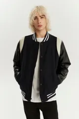 Campera estilo varsity con cuerpo de lana azul marino y mangas de cuero sintético en color negro. Presenta cuello, puños y ruedo acanalados con rayas blancas, cierre frontal con botones a presión y bolsillos laterales.