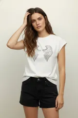 Remera blanca de algodón con cuello redondo y mangas cortas. Presenta un estampado de dos alas con apliques de strass plateados.