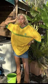 Remera amarilla de corte amplio con cuello en V y mangas cortas con ribete verde. Tiene dos líneas horizontales verdes en el pecho y el escudo de la selección de Brasil.