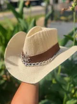 Sombrero de ala ancha tejido en hilo de coco y sintético, color marrón claro. La copa está cubierta con encaje naranja quemado con patrón floral. Tiene una cinta de cuero marrón oscuro y un aplique decorativo de hoja de gamuza con incrustaciones de pedrería.