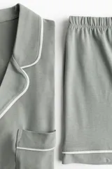Pijama corto de dos piezas color gris con ribetes blancos. Camisa de manga corta con cuello abierto, botones adelante, hombros caídos y bolsillo superior. Short con elástico en la cintura.