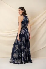 Vestido largo de fiesta en gasa plisada color azul con estampado plateado de hojas. Tiene falda amplia, cruzada y abierta, con bolsillos laterales. El escote es halter y tiene un cinto fino a la altura de la cintura.