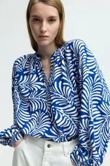 Blusa con estampado de hojas en azul y blanco, de tejido liviano, con cuello corto fruncido y tapeta de botones. Mangas globo largas con volumen de corte raglán y puños finos abotonados.