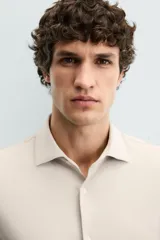 Camisa color beige de corte slim fit, confeccionada en tejido elástico con cuello italiano abierto, manga larga con puños abotonados y cierre frontal con botones.