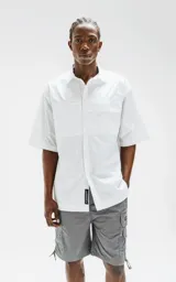 Camisa blanca de manga corta confeccionada en poplin de algodón, con silueta oversize y dos bolsillos delanteros, uno con solapa y otro con ojal y botón.