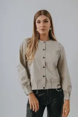 Campera bomber color beige de cuero, con cuello redondo, cierre frontal con botones a presión, mangas largas con puños elásticos y detalles de tachas metálicas en hombros y espalda.