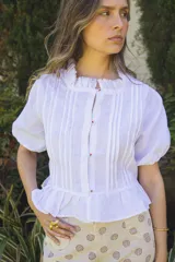 Camisa blanca de lino con alforzas verticales, cuello fruncido y mangas abullonadas con elástico. Los botones están bordados a mano con hilo de algodón. Tiene corte recto con pequeño volado en la base.