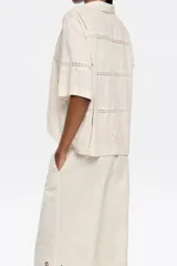 Camisa color crema de manga corta con cuello clásico y cierre frontal con lazos. Presenta bordados florales y calados.