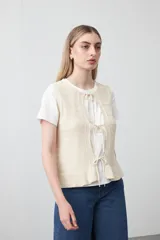 Chaleco tejido de punto color beige con trama texturada y diseño liviano. Presenta cuello a la base, silueta recta y cierre frontal mediante tres lazos delicados.