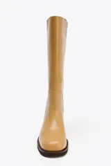 Bota alta de cuero color camel, con caña recta y punta redondeada. Presenta pespuntes decorativos verticales en la parte delantera y un tacón ancho de madera.