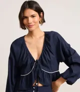 Blusa de mangas largas color azul marino, confeccionada en viscolino con 30% de lino, con escote V, lazo en el cuello con punta y bieses contrastantes luego abajo del escote, además del bajo de volado.