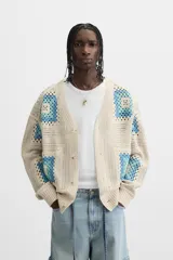 Cárdigan de punto color crudo con diseño de cuadrados de crochet en tonos celeste y azul. Tiene cuello en V, manga larga, bolsillos frontales y cierre con botones.