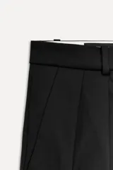 Pantalón negro de tiro alto con trabillas, bolsillos laterales y falsos bolsillos de vivo con solapa en la espalda. Detalle de cinturilla interior a contraste con pespuntes. Pinzas en el delantero. Cierre frontal con cremallera, botón interior y gancho metálico.