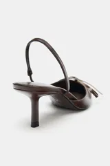 Zapato destalonado de mujer, color marrón oscuro con acabado brillante, punta fina y tacón alto y fino. Presenta un detalle de lazo con herraje dorado en la parte delantera y tira elástica en el talón.