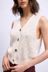 Chaleco de tejido de punto color crudo, sin mangas, con escote en V y cierre frontal de botones marrones.