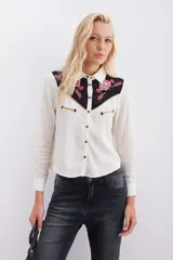 Camisa blanca de viscosa con canesú negro bordado con motivos florales en rosa y dorado. Tiene mangas largas con puños con snap metálico, acceso frontal con snaps metálicos y vivos en lurex dorado en canesú y en bolsillos ojales decorativos.
