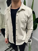 Chaqueta color beige de corte recto, con cuello camisero, cierre frontal con botones metálicos a presión, dos bolsillos con solapa abotonada en el pecho y mangas largas con puños abotonados.