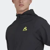 Cortavientos deportivo Le Coq Sportif, color negro, con capucha y cierre frontal. Presenta el logo del gallo en amarillo neón en el pecho izquierdo. El tejido es ligero y arrugado, típico de cortavientos.