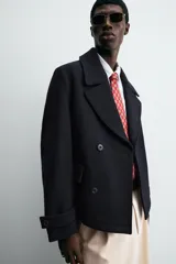 Abrigo corto azul marino de corte relajado, confeccionado en tejido con lana. Tiene cuello con solapas de pico, mangas largas con puños ajustables con trabilla, bolsillos de solapa en la cadera, bolsillo interior, abertura en el centro de la espalda y cierre frontal con botonadura.