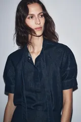 Camisa de denim azul oscuro con cuello redondo ajustable con lazadas, mangas cortas abullonadas, efecto lavado, bajo recto y cierre frontal con botones.