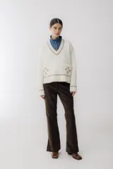 Sweater beige con cuello en V y mangas largas con diseño trenzado. Tiene dos bolsillos frontales con bordados de tréboles rojos y ribetes rojos en cuello, puños y ruedo.