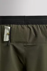 Shorts deportivos color verde oliva confeccionados en tejido técnico ligero y elástico. Cuenta con forro interior, cintura elástica ajustable con cordón, bolsillos laterales, bolsillo de seguridad en espalda con cierre de cremallera, bajo con aberturas laterales y logo estampado en la pierna.