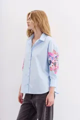 Camisa blanca de algodón con bordados florales y lentejuelas en las mangas, cuello camisero, mangas largas con puños abotonados y corte amplio.