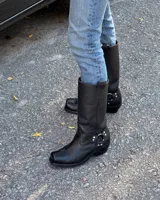 Bota alta de cuero vacuno color negro, con caña recta, puntera cuadrada y detalle de arnés con hebilla metálica plateada.