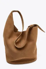 Bolso tipo saca asimétrica color marrón, confeccionado en serraje. Tiene asa bandolera ajustable y cierre con imán. Incluye una cartera con bolsillo extraíble.