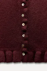 Cárdigan gris jaspeado de punto con cuello redondo y manga larga. Presenta volantes en el borde de los botones, en el bajo y en los puños.