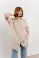 Camisa larga color beige de lino, con corte recto y calce suelto. Presenta cuello camisero, botones en el frente y mangas cortas que se pueden arremangar con presilla y botón.