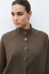 Chaqueta tejida de punto color verde oliva, con cuello alto y cierre frontal de botones a presión. Presenta bolsillos delanteros aplicados y mangas amplias de largo tres cuartos.