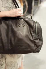 Bolso de viaje tipo duffel de cuero color marrón con doble asa de mano y correa bandolera ajustable y desmontable. Tiene cierre superior con cremallera y bolsillos laterales con cremallera.